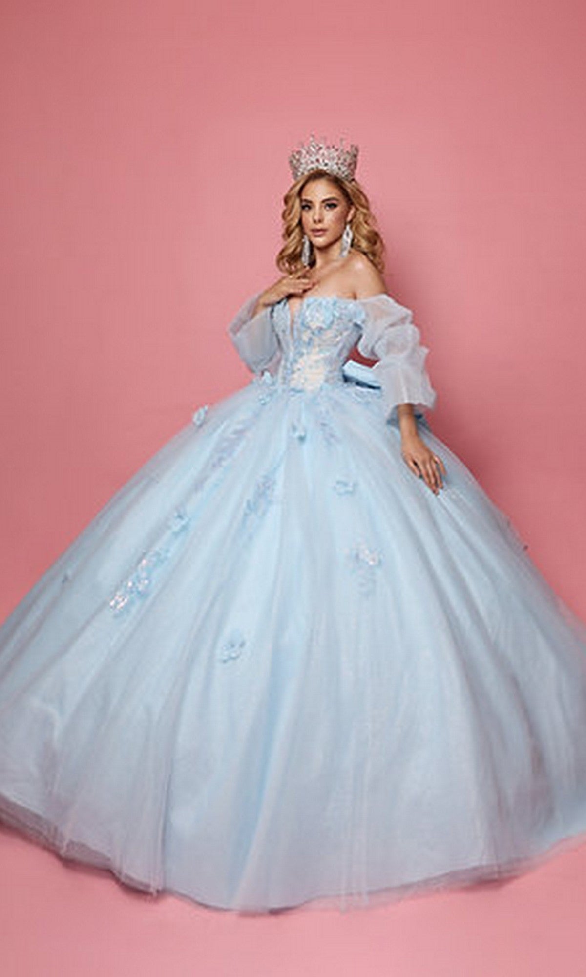 A woman stands against a pink background wearing the Quinceanera Dress Q121 By Nina Canacci in baby blue, featuring 3D floral appliques and an off-the-shoulder neckline, elegantly crowned.